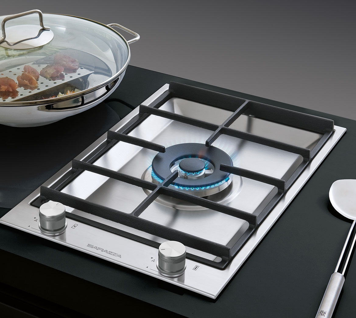 Dettaglio di un piano cottura a gas in acciaio inox satinato con un fuoco acceso, griglia in ghisa e due manopole per l'accensione, affiancato da un utensile e un wok contenente del cibo