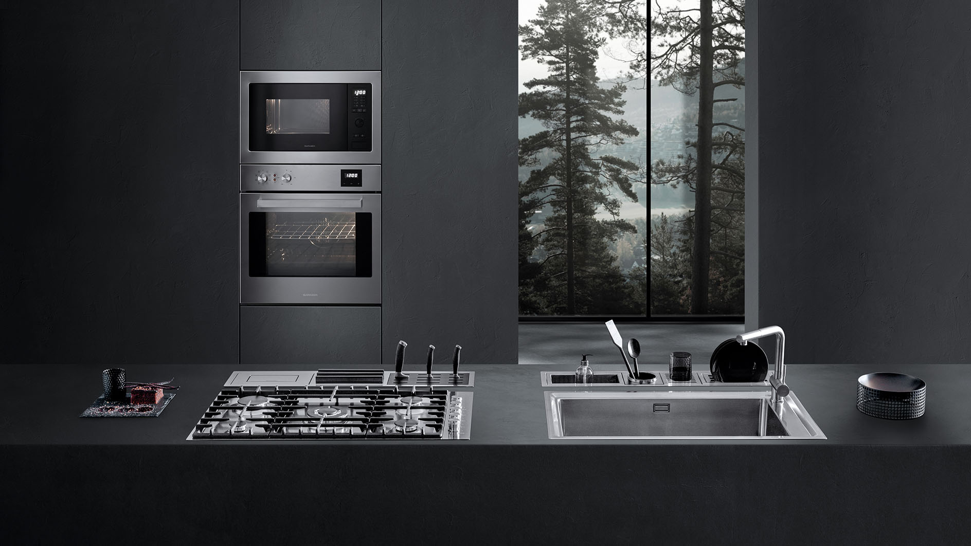 Vista di una cucina dotata di piano cottura a gas, lavello a vasca quadra, canali attrezzati con vari accessori e due forni a incasso. Sullo sfondo c'è una finestra con vista sul paesaggio esterno
