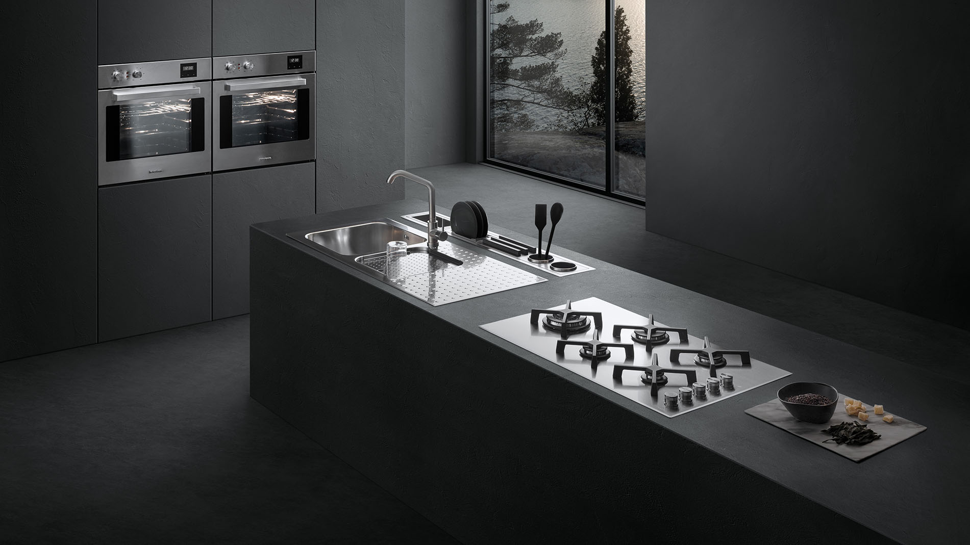 Vista di una cucina dotata di piano cottura a gas, lavello a vasca quadra con piano d'appoggio, un canale attrezzato con vari accessori e due forni a incasso. Sullo sfondo c'è una finestra con vista sul paesaggio esterno