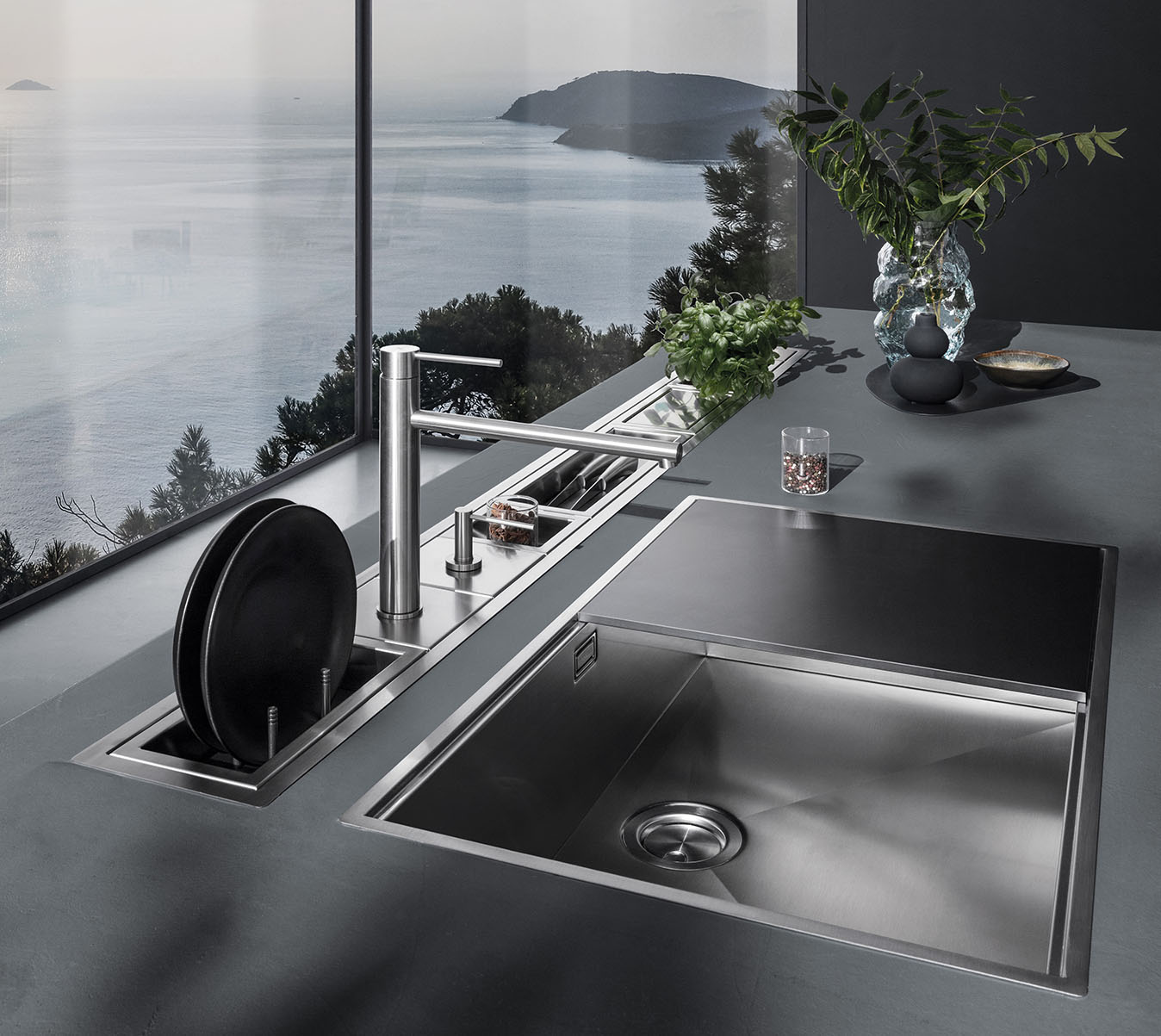 Dettaglio di una cucina in cui figurano un lavello in acciaio inox a vasca quadra con ripiano d'appoggio e un canale attrezzato in acciaio inox con svariati accessori. Sullo sfondo c'è una vetrata con vista sul paesaggio esterno