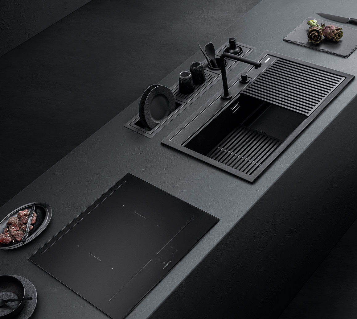 Visione di un piano da cucina nero su cui stanno affiancati un piano cottura a induzione e un lavello con sgocciolatoio. Sul piano di lavoro ci sono delle stoviglie e degli accessori.