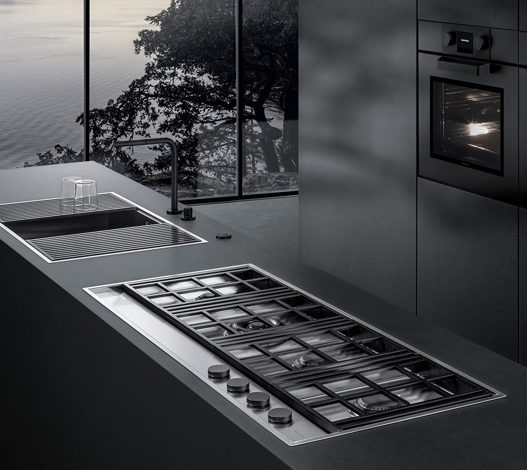 Vista di una cucina con forno sullo sfondo e, in primo piano, un piano cottura a gas che si affianca a un lavello con griglie di sgocciolamento.