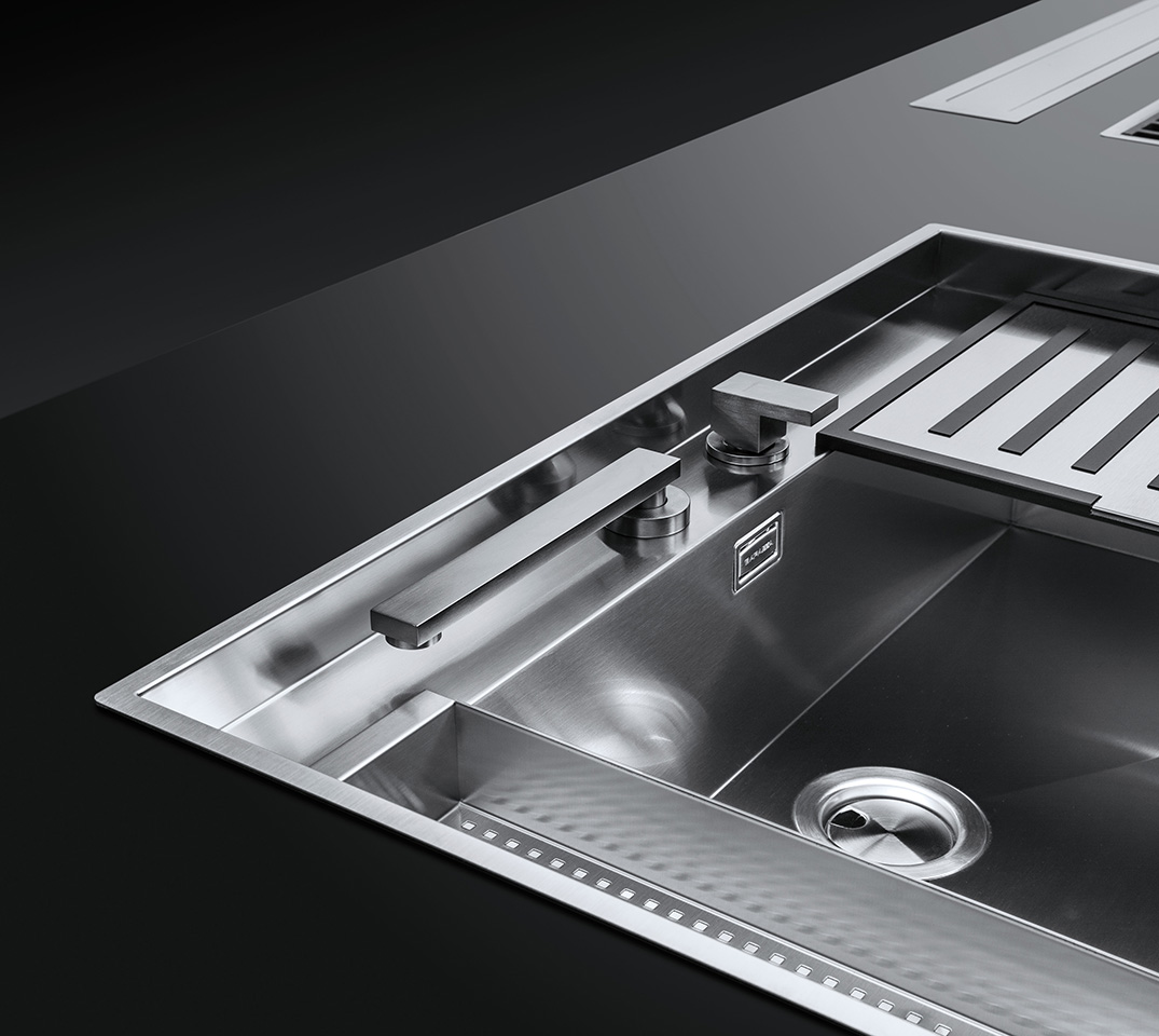 Dettaglio di un lavello in acciaio inox con vasca quadra, rubinetto miscelatore abbattibile e accessori.