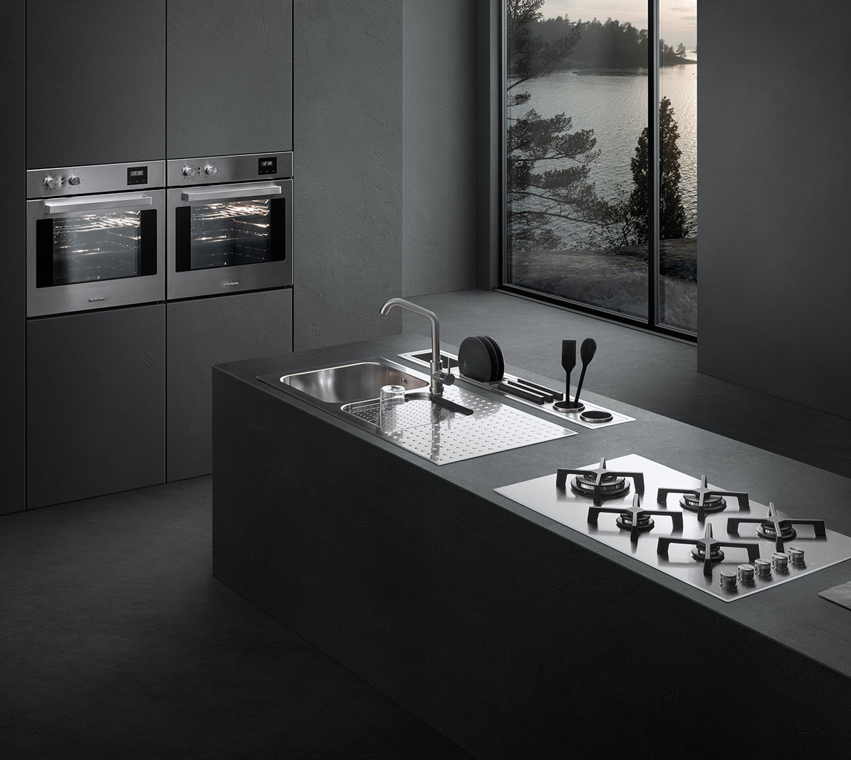 Vista di una cucina dotata di piano cottura a gas, lavello a vasca quadra con piano d'appoggio, un canale attrezzato con vari accessori e due forni a incasso. Sullo sfondo c'è una finestra con vista sul paesaggio esterno