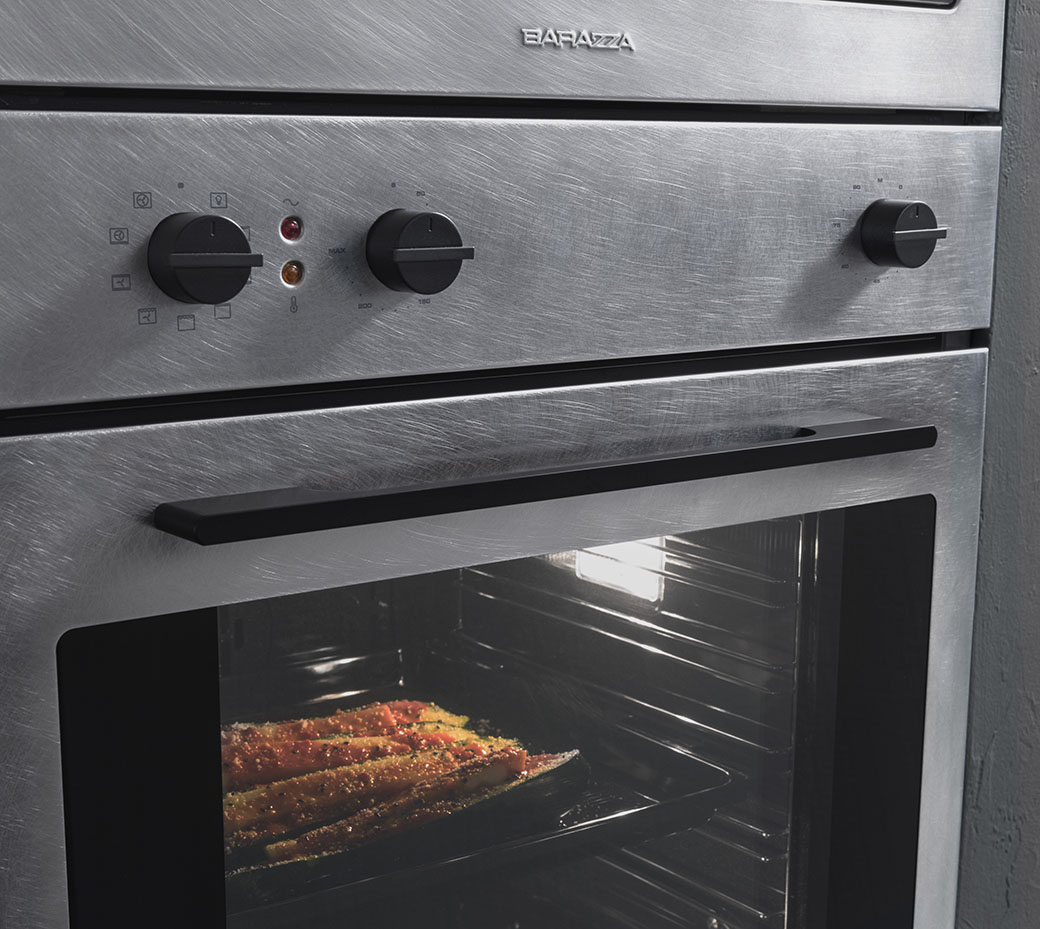 Dettaglio di un forno in acciaio inox finitura vintage con vetro frontale sulla porta, maniglione di apertura e tre manopole nere. Attraverso il vetro si può notare una teglia con delle verdure in cottura