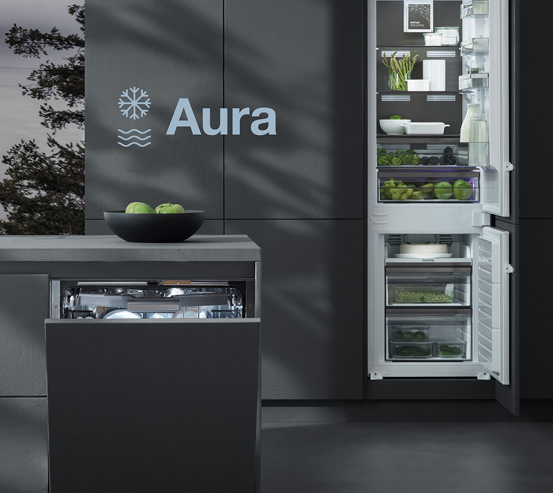 Barazza Assistenza Aura Dettaglio di una cucina in cui figurano un frigorifero a colonna con sportelli aperti e contenente frutta e verdura, una lavastoviglie semi aperta incassata in un piano su cui poggia una ciotola con delle mele. L'immagine reca la scritta "Aura" affiancata da due simboli