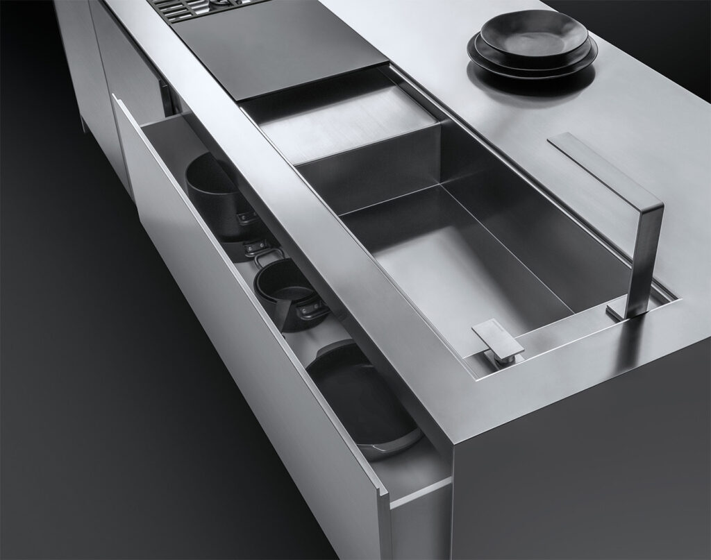 Dettaglio di un piano da cucina su cui figurano un lavello a vasca quadra con rubinetto e piano d'appoggio, parte di un piano di cottura a gas, una pila di piatti e un cassettone semi aperto contenente delle pentole