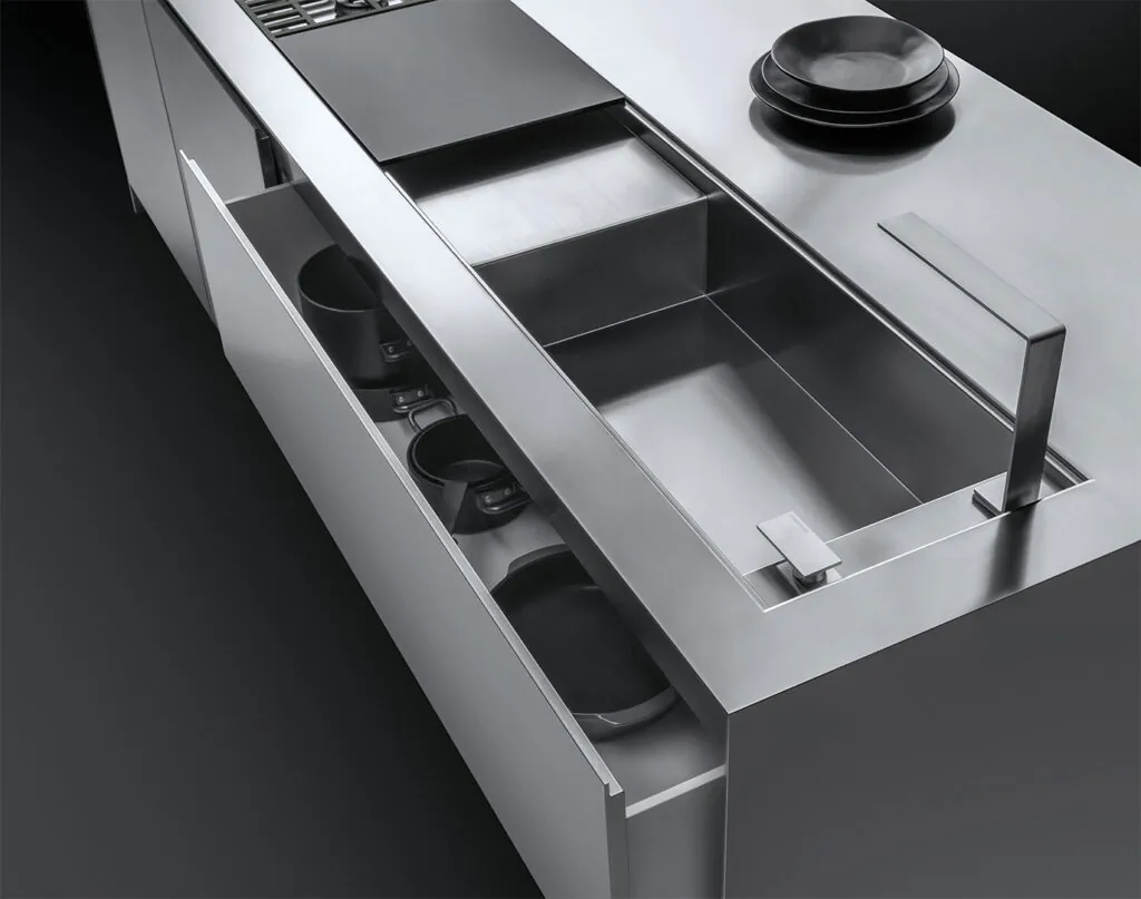 Dettaglio di un piano da cucina su cui figurano un lavello a vasca quadra con rubinetto e piano d'appoggio, parte di un piano di cottura a gas, una pila di piatti e un cassettone semi aperto contenente delle pentole