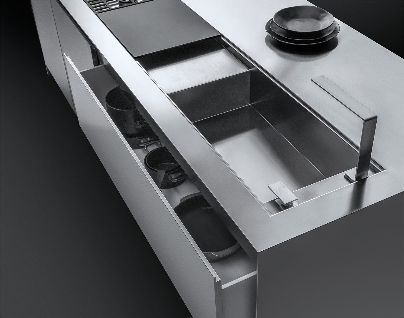 Dettaglio di un piano da cucina su cui figurano un lavello a vasca quadra con rubinetto e piano d'appoggio, parte di un piano di cottura a gas, una pila di piatti e un cassettone semi aperto contenente delle pentole