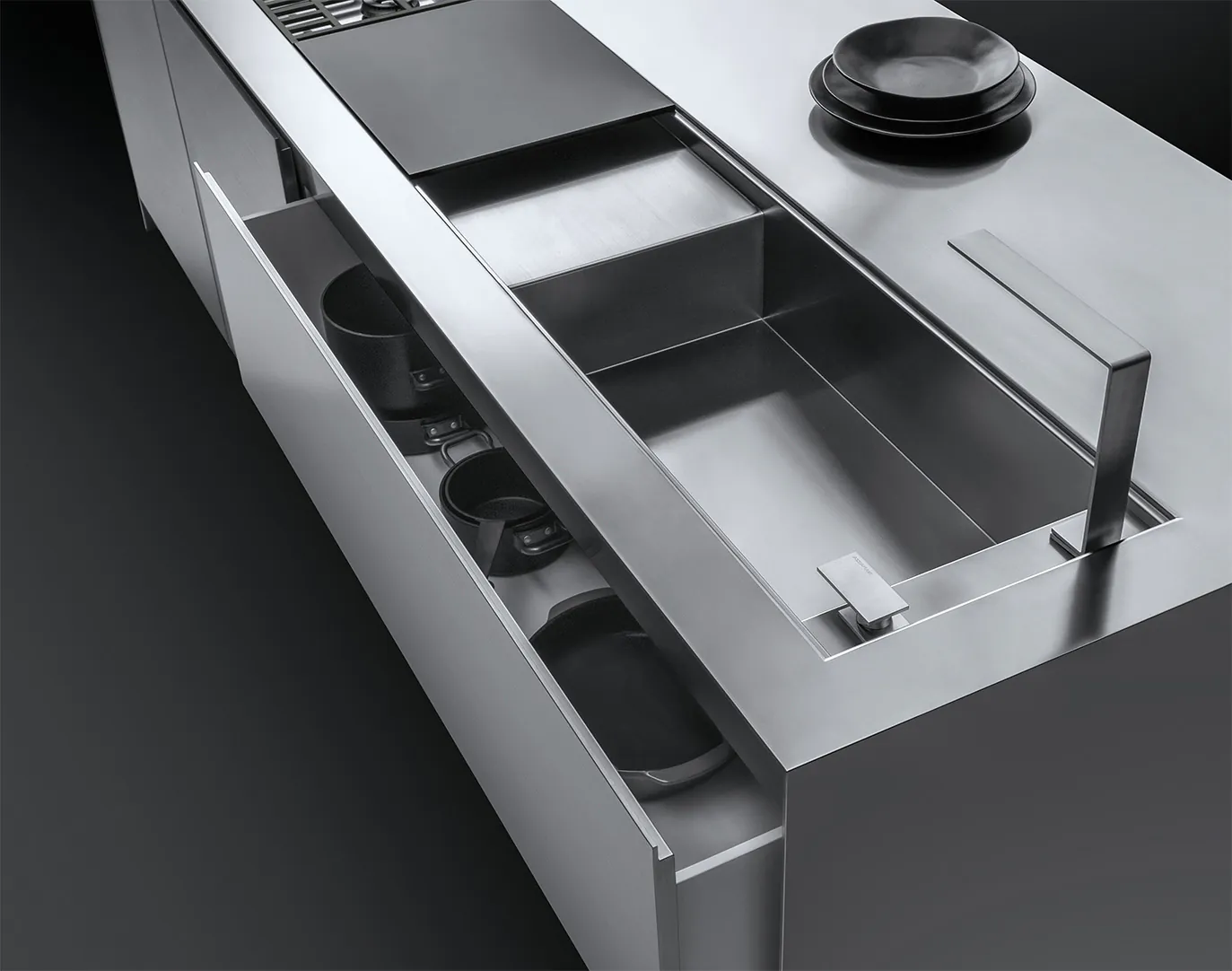 Dettaglio di un piano da cucina su cui figurano un lavello a vasca quadra con rubinetto e piano d'appoggio, parte di un piano di cottura a gas, una pila di piatti e un cassettone semi aperto contenente delle pentole