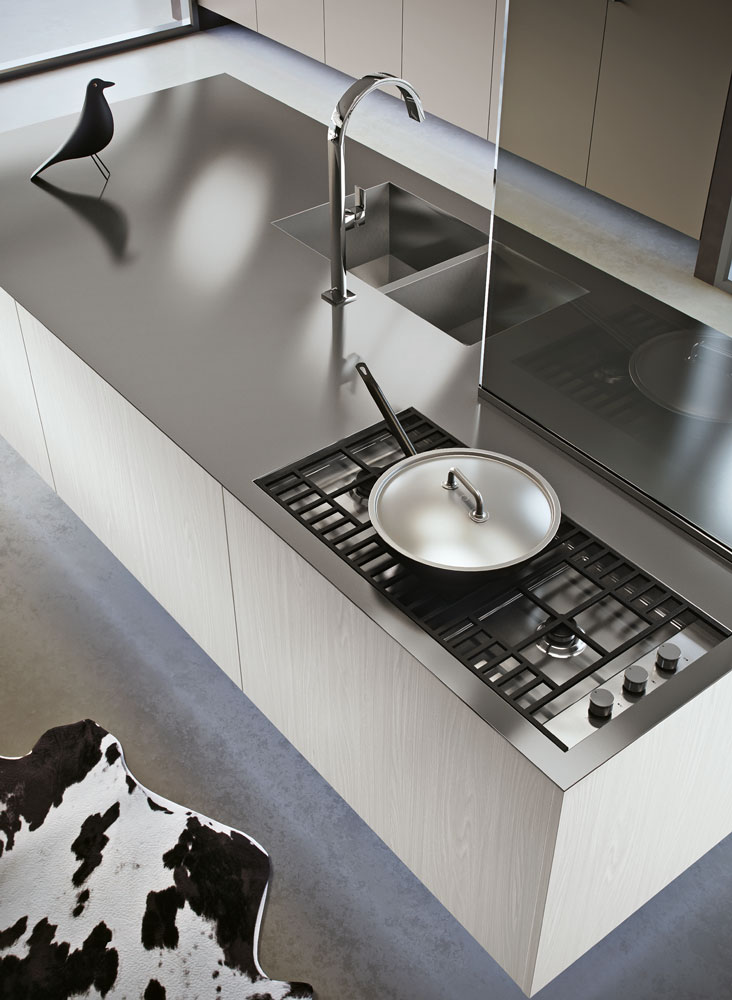 Dettaglio di una cucina in cui si vede un'isola in legno con piano in acciaio su cui figurano un piano di cottura a gas con sopra una padella e, posto di fronte separato da un divisorio in vetro, un lavello a due vasche quadre