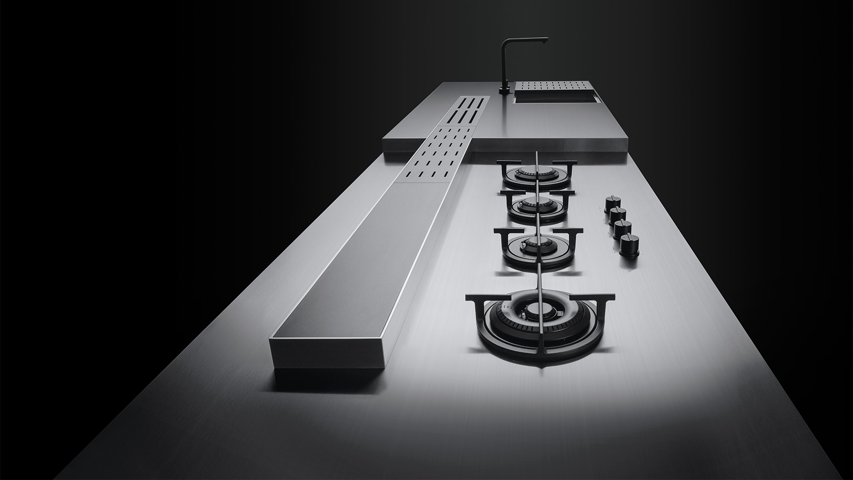 Vista longitudinale di un piano da cucina personalizzato sul quale spiccano dei fuori a gas in linea, un canale attrezzato e lavello con accessori.