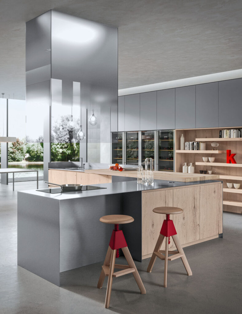 Vista di una cucina in legno e acciaio sul quale sfondo si nota una parete con scaffalature in legno su cui poggiano libri e oggetti di decoro affiancate da frigoriferi a parete. In centro figura un'isola angolare sormontata da una cappa e dei lampadiri, di fronte alla quale sono posti due sgabelli, sui cui poggiano un piano a induzione e un lavello