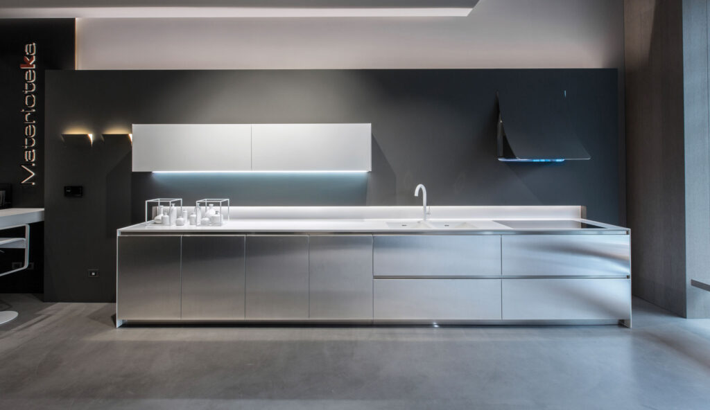 Vista frontale di una cucina a parete nella quale figurano un piano, montato su armadietti, su quale poggiano un lavello a due vasche quadre e un piano a induzione sormontato da una cappa aspirante.Sopra alla cucina sono montati degli stipetti a parete