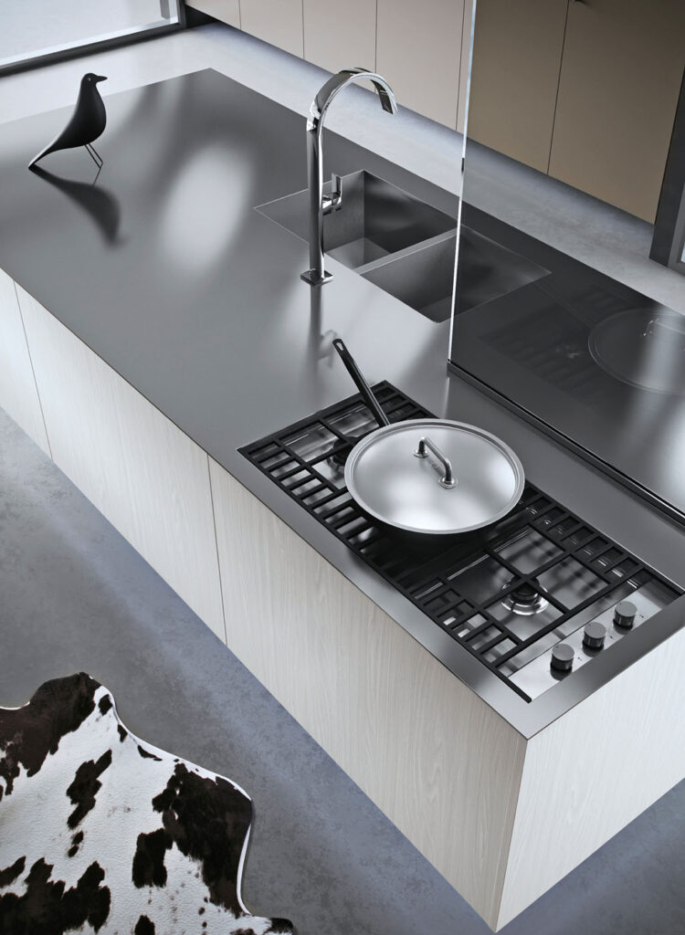 Dettaglio di una cucina in cui si vede un'isola in legno con piano in acciaio su cui figurano un piano di cottura a gas con sopra una padella e, posto di fronte separato da un divisorio in vetro, un lavello a due vasche quadre
