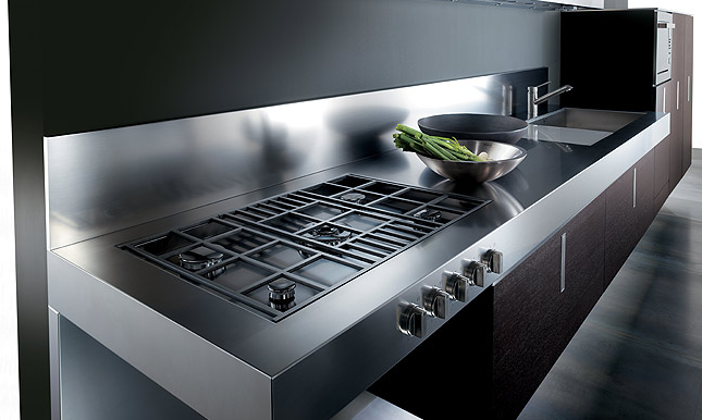 Dettaglio di una cucina con piano in acciaio sul quale si affiancano un piano cottura a gas a cinque fuochi e un lavello a vasca quadra.Di fianco al lavello c'è un forno incassato nel mobilio verticale