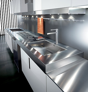 Cucina a parete dove figura un piano in acciaio montato su degli armadietti sul quale ci sono due lavelli a vasca quadra, posti a destra e a sinistra, in mezzo ai quali poggia un piano cottura a gas. Il piano è sormontato da una cappa aspirante