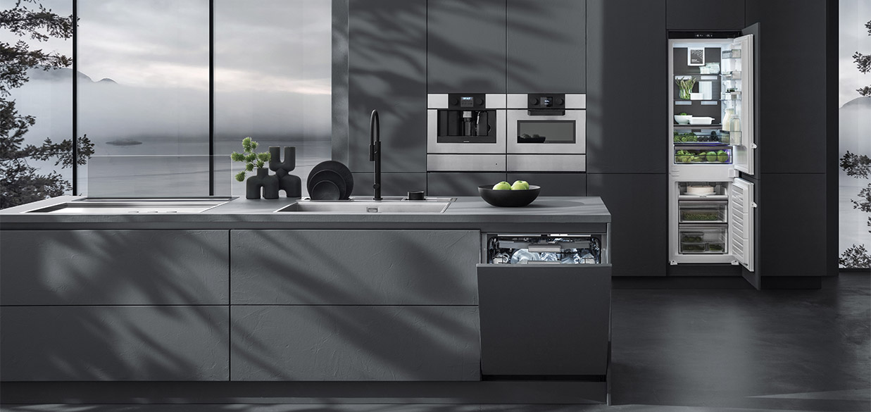 Vista di una cucina dotata di piano cottura a induzione con cappa aspirante a incasso, lavello a vasca quadra, un canale attrezzato con vari accessori, due forni a incasso, frigorifero a colonna e lavastoviglie a incasso. Sullo sfondo c'è una finestra con vista sul paesaggio esterno.
