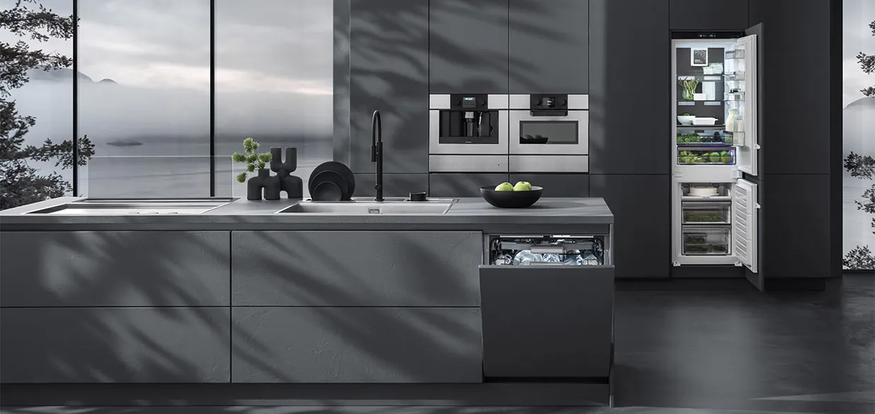 Vista di una cucina dotata di piano cottura a induzione con cappa aspirante a incasso, lavello a vasca quadra, un canale attrezzato con vari accessori, due forni a incasso, frigorifero a colonna e lavastoviglie a incasso. Sullo sfondo c'è una finestra con vista sul paesaggio esterno.