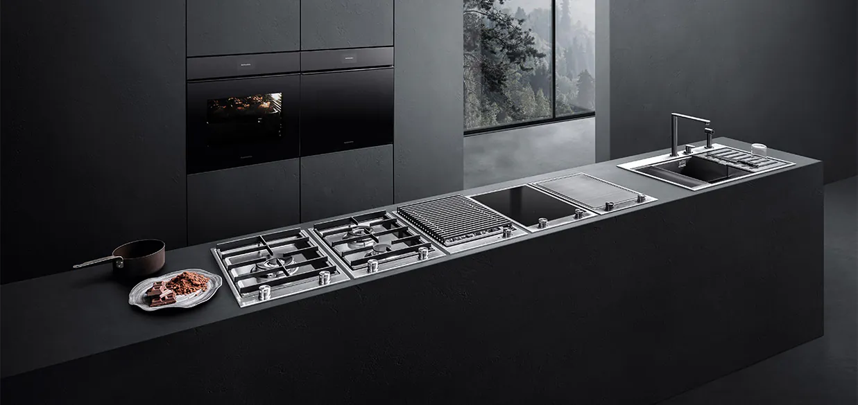 Vista di una cucina dotata di vari piani cottura, lavello a vasca quadra con accessori e due forni a incasso. Sullo sfondo c'è una finestra con vista sul paesaggio esterno.