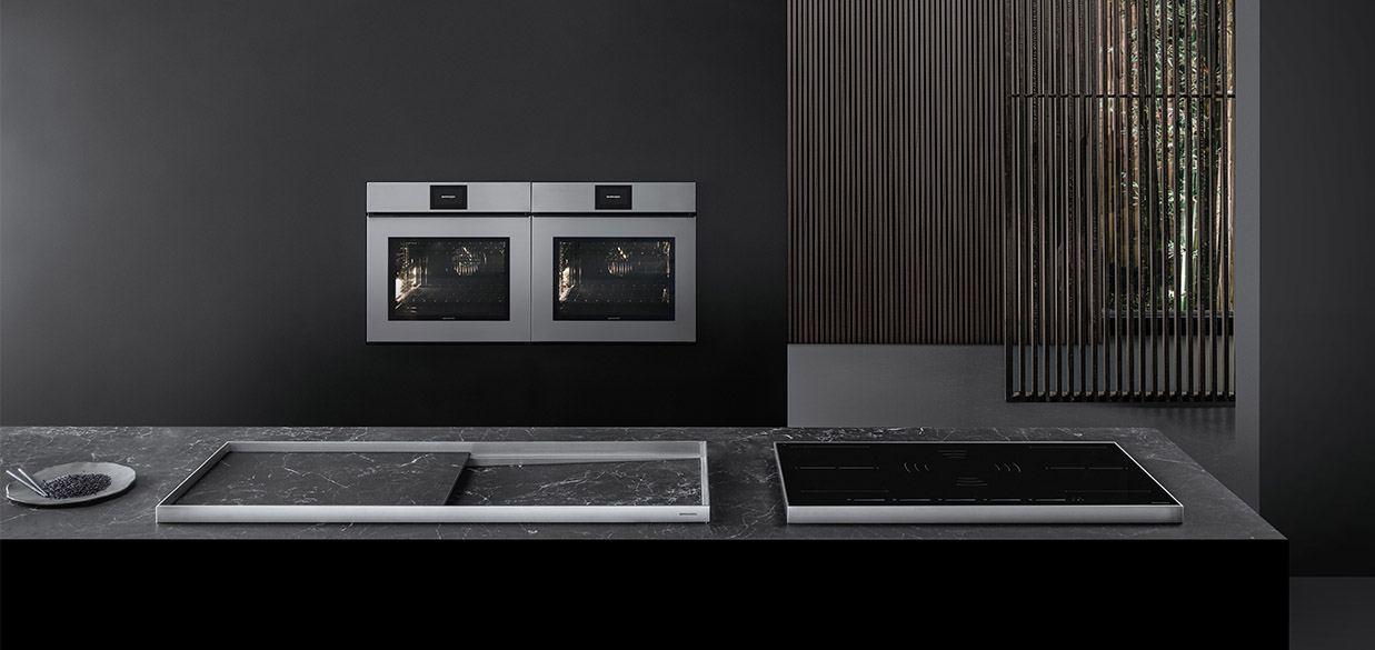 Vista di una cucina dotata di piano cottura a induzione, lavello a vasca quadra con piano d'appoggio, e due forni a incasso. Sullo sfondo c'è una stanza con divisori in legno.