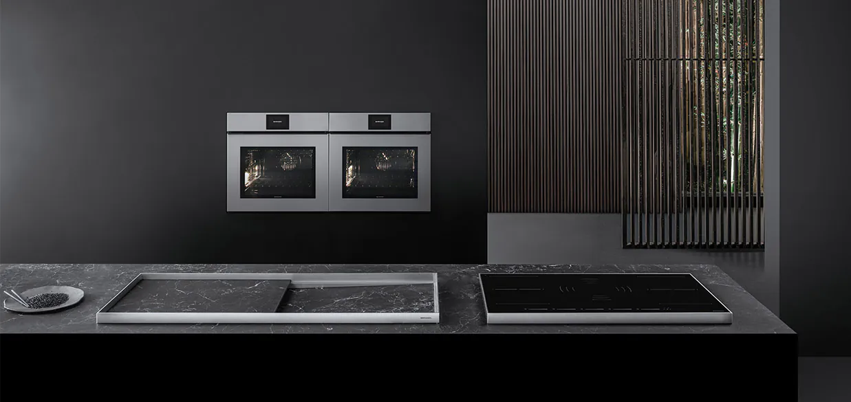 Vista di una cucina dotata di piano cottura a induzione, lavello a vasca quadra con piano d'appoggio, e due forni a incasso. Sullo sfondo c'è una stanza con divisori in legno.