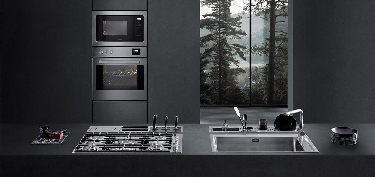 Vista di una cucina dotata di piano cottura a gas, lavello a vasca quadra, canali attrezzati con vari accessori e due forni a incasso. Sullo sfondo c'è una finestra con vista sul paesaggio esterno.