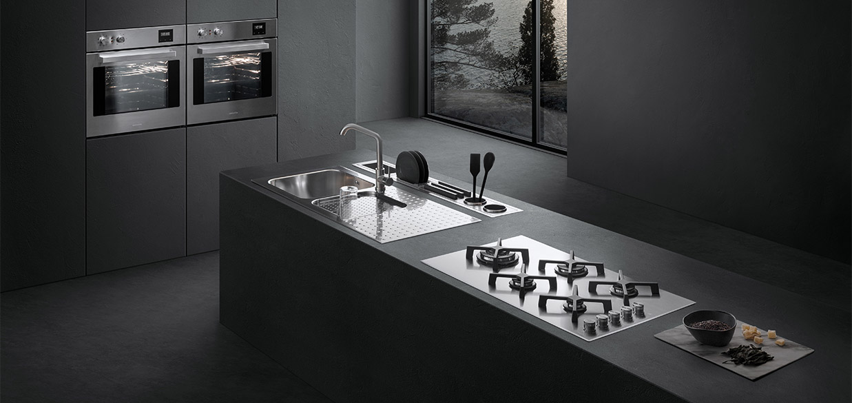 Vista di una cucina dotata di piano cottura a gas, lavello a vasca quadra con piano d'appoggio, un canale attrezzato con vari accessori e due forni a incasso. Sullo sfondo c'è una finestra con vista sul paesaggio esterno.