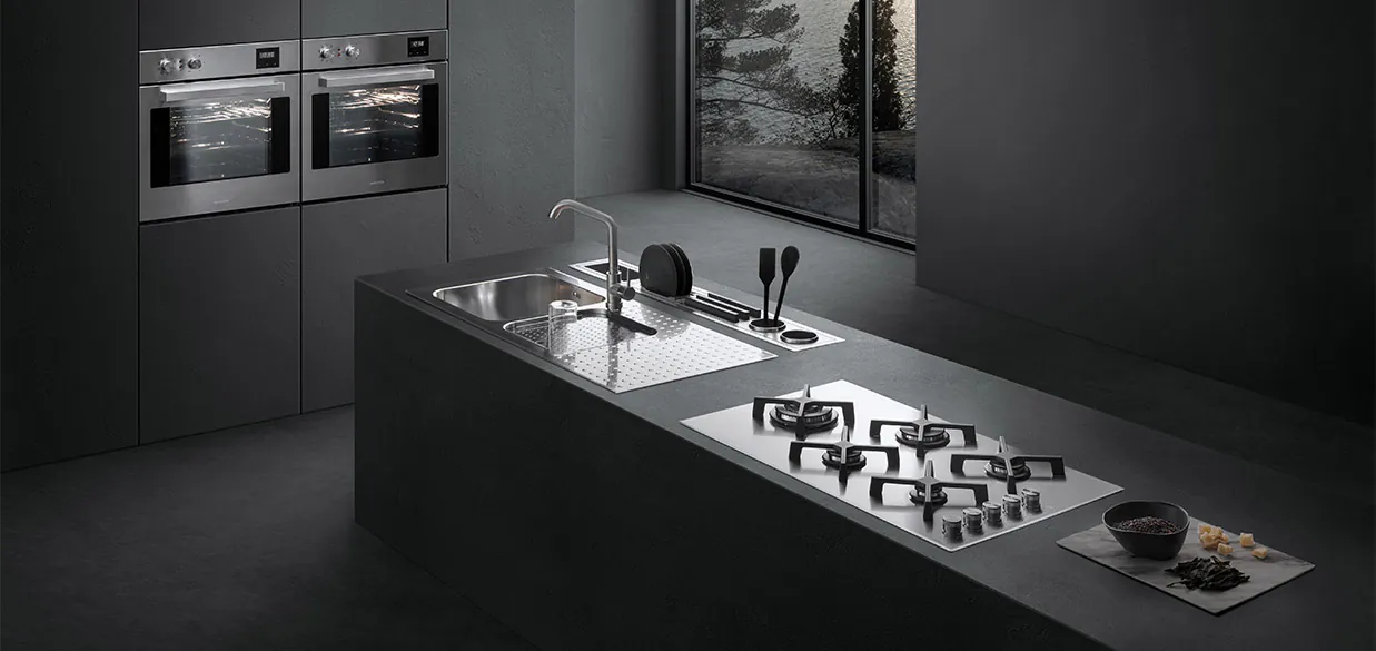 Vista di una cucina dotata di piano cottura a gas, lavello a vasca quadra con piano d'appoggio, un canale attrezzato con vari accessori e due forni a incasso. Sullo sfondo c'è una finestra con vista sul paesaggio esterno.