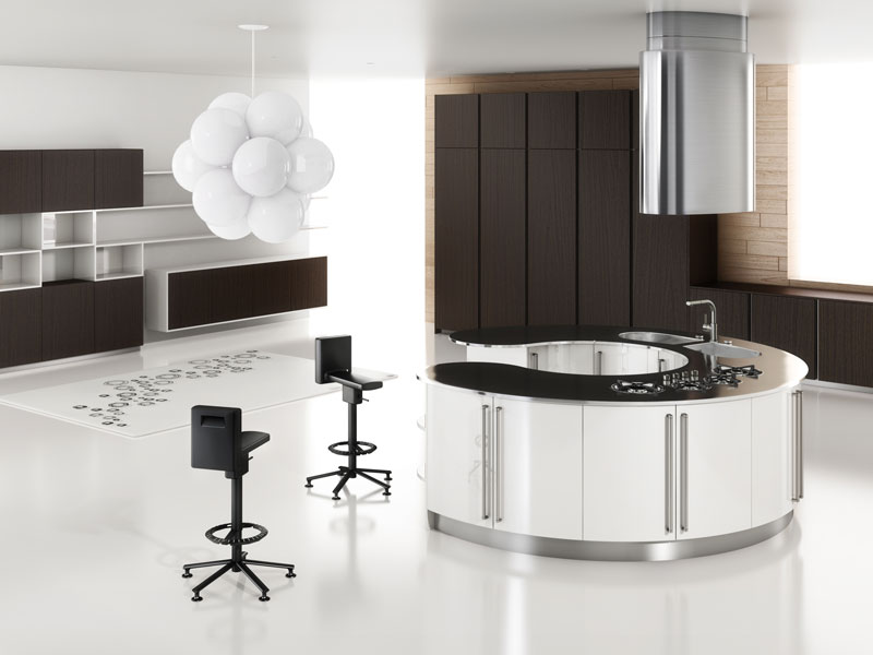 Vista di una zona living al cui centro figura un piano da cucina incurvato su cui si affiancano piani cottura a gas e un lavello a due vasche. Sullo sfondo si vedono una parete attrezzata e del mobilio verticale, di fianco al piano cucina ci sono due sedie