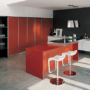 Vista di una cucina composta da un piano arancione e uno bianco che supportano un piano cottura in acciaio. 
In primo piano ci sono degli sgabelli posti di fronte al piano arancione e sullo sfondo delle scaffalature contenenti dei bicchieri