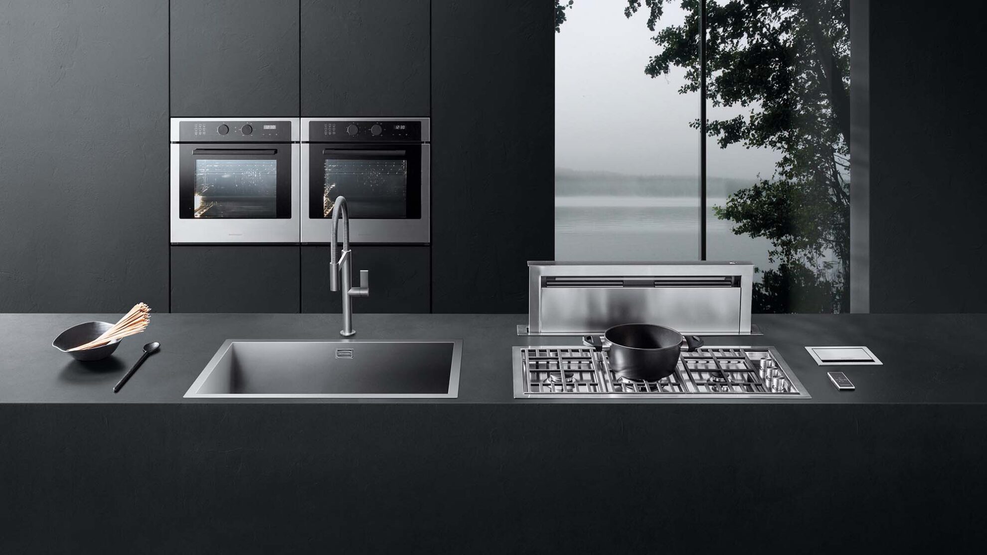 Cucina Barazza con lavello, rubinetto e piano cottura in primo piano, due forni sullo sfondo e vetrata laterale che mostra un albero all’esterno
