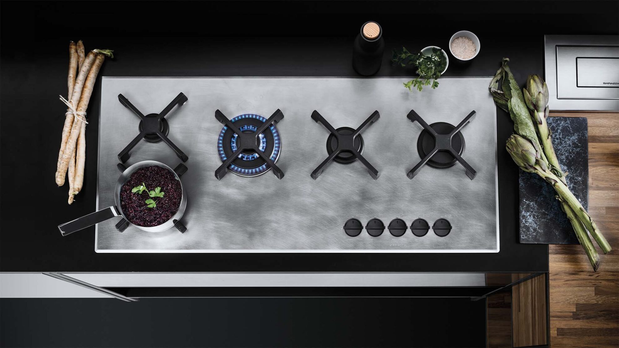 Piano cottura grigio della Collezione Unique con 5 fornelli, uno acceso, circondato da verdure fresche come carciofi e rapa, ingredienti base come sale e prezzemolo e una bottiglia nera decorativa
